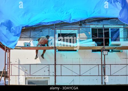 Unbekannter Arbeiter, der auf einem Gerüst sitzt und Wärmedämmung aus Schaumplatten an der Hauswand installiert Stockfoto