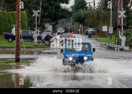 Burlington, Washington, USA. Dezember 2025. Die Autos fahren am Freitag, den 12. Dezember, durch das Wasser von den überfluteten Gages Slough, einem Nebenfluss des Skagit River, an der Kreuzung von Gardner Road und State Route 20 in Burlington, Washington, USA. 2025, nachdem drei atmosphärische Flüsse die Region durchnässt hatten. Früh am Morgen befahlen die Behörden den Einwohnern, die Stadt zu evakuieren, da das Hochwasser anfing zu steigen. Prognosen zufolge würden viele Flüsse im pazifischen Nordwesten katastrophale Hochwasserstände erreichen. Der Gouverneur hat den Ausnahmezustand ausgerufen und heute hat die US-Regierung die Überschwemmung ausgerufen Stockfoto