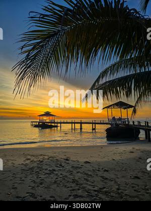 Goldene Farbtöne erfüllen den Himmel, während die Sonne über Koh Kood Island, Thailand, untergeht. Ein ruhiger Pier erstreckt sich in das ruhige Wasser, eingerahmt von üppigen Palmen, die ein friedliches Paradies für Entspannung schaffen. Stockfoto
