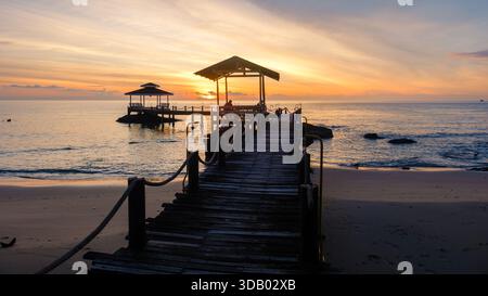 Goldene Farbtöne erleuchten den Himmel, während die Sonne über dem ruhigen Wasser der Insel Koh Kood Thailand untergeht. Der rustikale Holzsteg erstreckt sich bis in den Horizont und lädt zu abendlichen Spaziergängen und friedlichen Reflexionen ein. Stockfoto