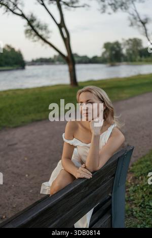 Lächelnde Frau in weißem Kleid, die auf einer modernen Holzbank im Stadtpark sitzt Stockfoto
