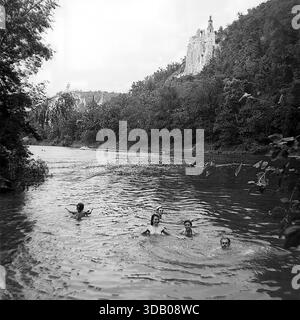 Slavyanogorsk (Svyatogorsk), Ukraine, UdSSR, 1950er Jahre Ein malerisches Schwarzweiß-Archiv, das die Sommerferien auf dem Seversky Donets River aufnimmt. Eine Gruppe glücklicher sowjetischer Leute schwimmt und badet im ruhigen Wasser. Im Hintergrund, die majestätischen Kreidefelsen und die historische St. Nikolaus Kirche der Heiligen Berge Lavra Turm über dem Fluss. Dieses stimmungsvolle Bild veranschaulicht anschaulich die Schönheit der Natur und den unbeschwerten Urlaub in der friedlichen Donbass-Region während der Sowjetzeit. Stockfoto