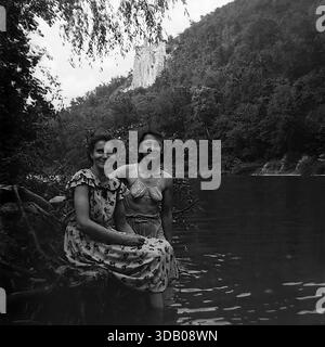 Slavyanogorsk (Svyatogorsk), Ukraine, UdSSR, 1950er Jahre Ein bezauberndes Schwarz-weiß-Archivfoto, das zwei glückliche sowjetische Frauen zeigt, die auf Baumwurzeln am Ufer des Seversky Donets sitzen. Der eine trägt ein Blumenkleid, der andere ein Badeanzug-Oberteil. Im Hintergrund die majestätischen Kreidefelsen und der historische Nikolaikirche über dem Wasser. Dieses Bild zeigt anschaulich weibliche Freundschaft, Sommermode und ruhige Freizeit im friedlichen Donbass. Stockfoto