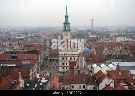 Hoher symmetrischer Blick auf den Rathausturm von Poznań und den Altstadtplatz, rote Ziegeldächer und Straßen, entfernte Stadt mit Dunst, grauer Himmel. Stockfoto