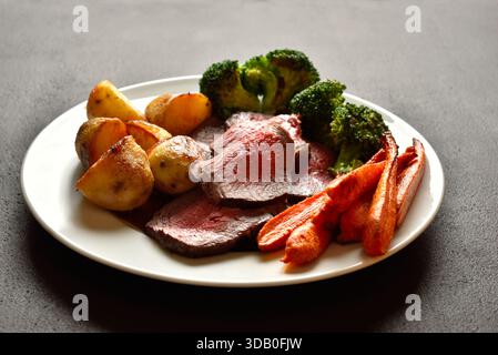 Sonntagsbraten mit gebratenem Rindfleisch, Karotten, knuspriger Kartoffel und Brokkoli auf weißer Platte auf braunem Steinhintergrund. Traditionelles britisches Gericht. Nahansicht Stockfoto