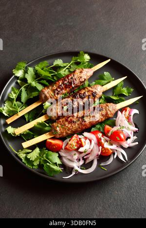 Nahansicht von lula Kebab auf Holzspießen serviert mit frischem Gemüse auf schwarzem Teller auf dunklem Hintergrund. Stockfoto