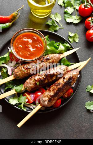 Gegrillter lula-Kebab auf Holzspießen serviert mit frischem Gemüse und Tomatensauce auf schwarzem Teller auf dunklem Hintergrund. Nahansicht Stockfoto