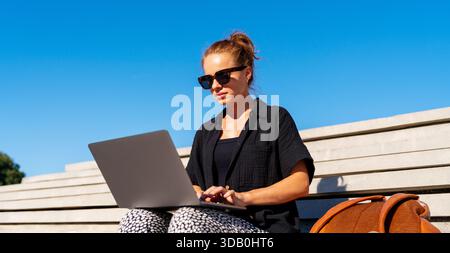 Selbstbewusste Frau mit Laptop auf urbanen Stufen in der Stadt vor blauem Himmel. Stockfoto
