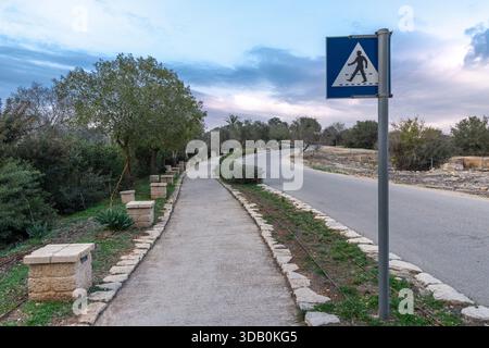 Kurvenreiche Straße und Fußweg mit einem Fußgängerschild im Zippori-Nationalpark im Norden Israels. Ruhiges Abendlicht, Olivenbäume und bewölkter Himmel sorgen für eine ruhige Atmosphäre Stockfoto