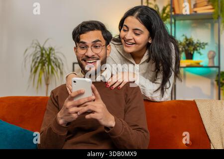 Lächelnde indische Frau, die mit einem Freund spricht, der das Smartphone durchzieht und zu Hause auf dem Sofa sitzt. Multiethnisches junges Paar, das zusammen ein Mobiltelefon benutzt, um online Lieferung in Wohnzimmerwohnung zu bestellen. Stockfoto