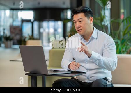 Junger chinesischer Geschäftsmann, der sich in der Hotellobby auf einem Laptop aufschreibt oder Notizen macht, während er an einem Online-Videokurs oder einem Büromeeting teilnimmt. Selbstbewusster Manager im Bereich T-Shirt-Support, der Webcam ansieht Stockfoto