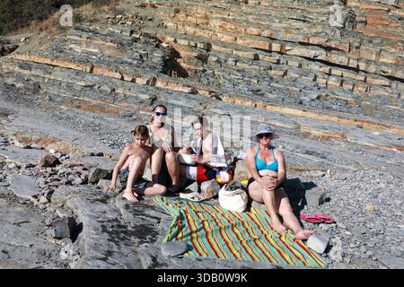 Zwei Frauen, ein junger Mann und ein Kind sitzen an einem felsigen Ufer vor geschichteten Felsformationen. Familienurlaub am Strand mit natürlicher Geologie. Stockfoto