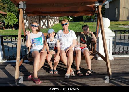 Frau mit zwei Jungen und einer älteren Frau, die auf einer Holzschaukel sitzt und gemeinsam den Sommerurlaub in einem Resort genießt. Familienverbundenheit und Freizeitkonzept. Stockfoto
