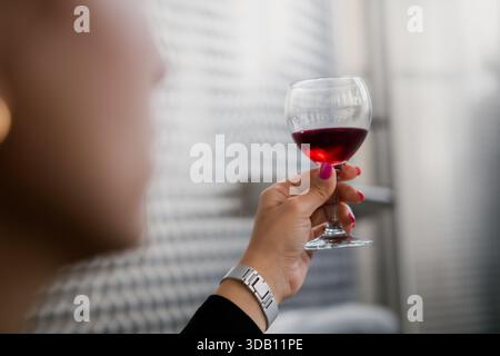 Die Hand hält ein lebendiges Rotweinglas. Verschwommener Hintergrund der Weinfabrik deutet auf Produktion oder Verkostung hin. Konzentrieren Sie sich auf die Farbe und Klarheit des Weins. Stockfoto