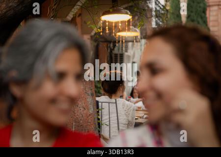 Tiflis, Georgien - 3 30 2025: Zwei verschwommene Frauen im Vordergrund bilden eine lebendige Café-Szene im Freien mit Hängeleuchten und Sitzplätzen. Stockfoto