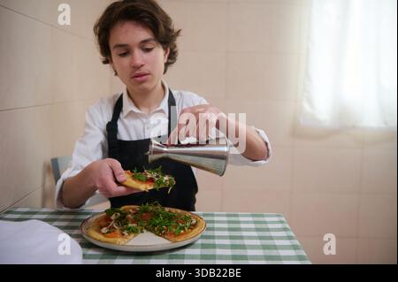 Ein junger Koch in weißem Hemd und Schürze gießt in einer gemütlichen Küche Sauce über eine frisch belegte Pizza. Stockfoto