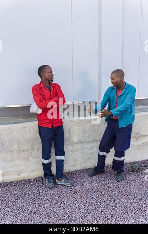 Zwei junge afrikanische Arbeiter, Arbeitskleidung, reden draußen neben einer Lagerwand Stockfoto