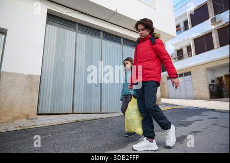 Zwei Kinder in Jacken laufen auf einer sonnigen Straße entlang und tragen eine gelbe Plastiktüte. Eine einfache Szene des Alltags und Familienausflugs in einer Wohnung Stockfoto