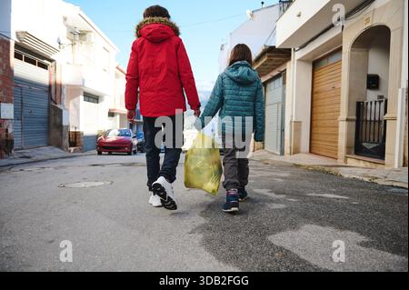 Zwei Kinder laufen Hand in Hand auf einer ruhigen Stadtstraße mit einer gelben Tasche. Sie tragen Winterjacken, was auf einen lässigen, alltäglichen Familienmoment hindeutet Stockfoto
