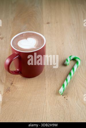 Ein roter Becher mit heißer Schokolade mit einem Herzen darauf sitzt auf einem Holztisch. Ein grün-weiß gestreifter Zuckerrohr ist neben dem Becher Stockfoto