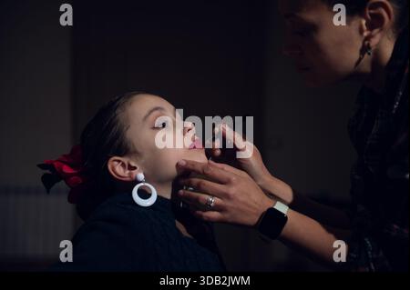 Ein fürsorglicher Moment, als eine Mutter ihren roten Lippenstift auf ihre Tochter aufträgt. Nahaufnahme-Make-up-Szene, die Vertrauen, Stil und Bindung in einer warmen, intimen Umgebung festhält. Stockfoto