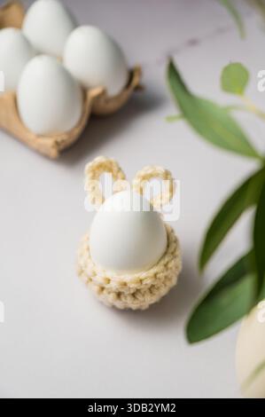 Osterhintergrund. Grüner Eukalyptus, weißes Ei in einem gestrickten Hasen-Eierhalter. Frühlingszeit. Minimalistisch. Stockfoto