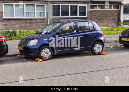 Ein geparktes blaues Auto, das durch zwei gelbe und rote Radklammern an den Reifen wegen eines Parkverstoßes in einer niederländischen Stadtstraße stillgelegt wurde. Stockfoto