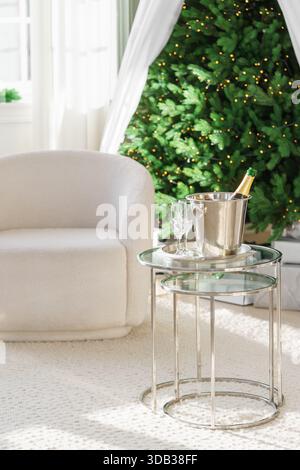 Champagnerflasche in Metall Eiskübel mit zwei leeren Gläsern auf Tisch im Wohnzimmer mit weißem Sessel auf glühendem Weihnachtsbaum Lichter Hintergrund. F Stockfoto
