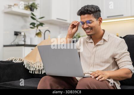Ein indischer Mann zu Hause hat Schwierigkeiten, winzige Texte auf dem Laptop zu lesen, zieht dann eine Brille auf und sieht klare Sicht. Arabian Guy on Sofa Lächeln passt den Rahmen an, liest Bildschirmlinien ohne Belastung und setzt die Arbeit fort. Stockfoto