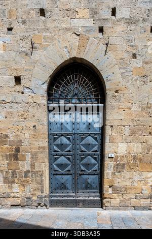 Mittelalterliches Steinportal des Palazzo dei priori in Volterra, stabile Eisentür unter spitzem Bogen auf der sonnendurchfluteten piazza. Stockfoto