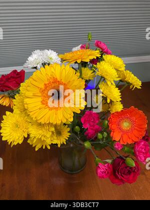 Ein Blumenstrauß mit gelben und orangefarbenen Gerbera Daisies, roten Rosen, rosa Nelken und gelben und weißen Chrysanthemen, in einer Vase, auf einem Küchentisch Stockfoto