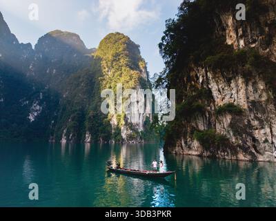 Ein Paar vor einem Langboot bei Sonnenaufgang auf dem Cheow Lan Lake in Khao Sok, Thailand. Das smaragdgrüne Wasser ist von hohen Kalksteinklippen umgeben und bietet eine perfekte Kulisse für Abenteuer Stockfoto