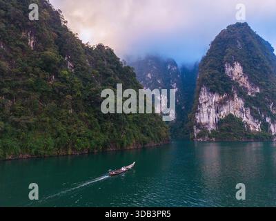 Erkunden Sie das ruhige Wasser des Cheow Lan Lake in Khao Sok, Thailand, während ein Holzboot an dramatischen Kalksteinklippen vorbeigleitet. Eine malerische Szene in der Abenddämmerung, die Natur und Abenteuer miteinander verbindet. Stockfoto