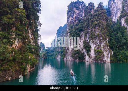 Eine ruhige Szene entfaltet sich, während ein kleines Boot über das türkisfarbene Wasser des Cheow Lan Lake gleitet, eingerahmt von hohen Kalksteinklippen. Khao Sok Thailand Stockfoto