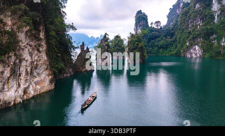Entdecken Sie die atemberaubende Schönheit des Cheow Lan Lake, wo das smaragdgrüne Wasser aufragende Kalksteinklippen reflektiert. Ein traditionelles Boot gleitet friedlich durch die ruhige Landschaft, Khao Sok Thailand Stockfoto