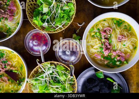 Schüsseln mit authentischer vietnamesischer Rindfleischnudelsuppe (Pho), garniert mit frischen Kräutern, Sojasprossen und Chili-Sauce auf einem rustikalen Holztisch Stockfoto