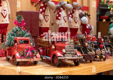 Auf einem Holzregal mit kleinen Weihnachtsbäumen und Bändern befindet sich eine bezaubernde Sammlung roter Vintage-Spielzeugwagen. Stockfoto