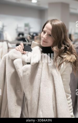Eine schöne junge, schöne Frau von 30-35 Jahren probiert einen stilvollen weißen Pelzmantel mit kurzem Pelz in einer stilvollen Mode-Boutique an. Festlicher Herbstwinter Stockfoto