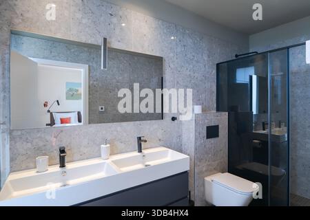 Modernes, luxuriöses Badezimmer mit zwei Waschbecken, eleganten grauen Marmorfliesen, ebenerdiger Dusche und minimalistischer Toilette. Ein Spiegel reflektiert einen Blick Stockfoto
