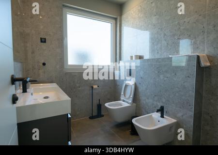 Modernes, luxuriöses Badezimmer mit zwei Waschbecken, eleganten grauen Marmorfliesen, ebenerdiger Dusche und minimalistischer Toilette. Ein Spiegel reflektiert einen Blick Stockfoto