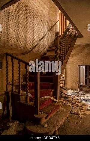 Verstaubte geschwungene Treppe mit rotem Teppich in einer verlassenen Villa mit verzierten, verdrehten Balustern und verstreuten Trümmern Stockfoto
