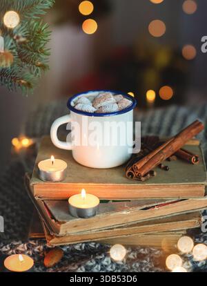 Gemütliche Winterszene mit einem weißen Becher gefüllt mit Marshmallows auf gestapelten Büchern, umgeben von Kerzen und Zimtstangen, die Wärme und Urlaub wecken Stockfoto