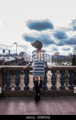 Eine Frau, die von hinten gesehen wird, steht auf der Fußgängerbrücke über den Main in Frankfurt und trägt ein schickes einteiliges Schwarz-weiß-Outfit. Stockfoto