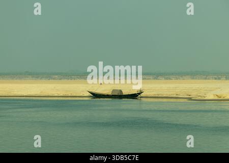 Holzfischboot vor Anker an einem Sandufer oder Flussufer mit einer kleinen zentralen Kabine und einer dunklen Figur auf dem Bug, die sich in ruhigem Blaugrün spiegelt Stockfoto
