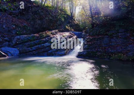 Ruhiger Waldwasserfall, der in sanftem Sonnenlicht über felsige Lagen fließt. Ein ruhiger kleiner Wasserfall, der über geschichtete Felsformationen in einem friedlichen F Stockfoto