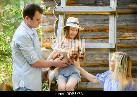 Familie genießt Urlaub im Freien mit Haustier. Mädchen sitzt im Schatten des Hauses an heißen Sommertagen und hält Hund. Ihre Eltern streicheln Spaniel, während sie mit ihrer Tochter spielen, und Haustiere im Hof des Landhauses Stockfoto