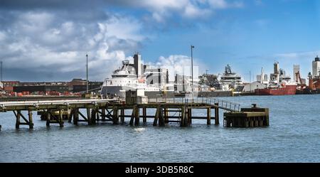 Ein geschäftiger Hafen mit einem verwitterten Pier, der sich über ruhiges Wasser erstreckt, große Schiffe und Industrieschiffe, die an den Anlegestellen unter einem dramatischen blauen Himmel angedockt sind. Stockfoto
