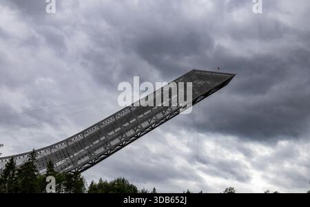 Oslo, Norwegen, 11. August 2025: Die legendäre Holmenkollbakken-Schanze erstreckt sich in den Himmel mit dramatischen grauen Wolken und Wald an der Basis. Stockfoto