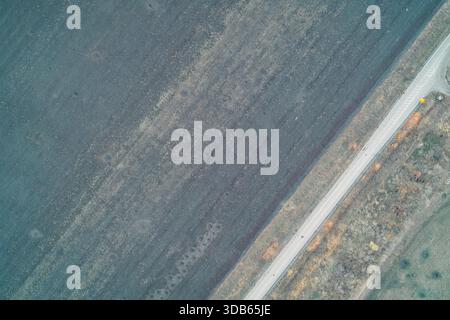 Von oben nach unten auf eine diagonale Asphaltstraße, die ein dunkles, verkratertes Ackerfeld von einem Waldgürtel trennt. Der Kontrast zwischen dem geschädigten Boden und der Verkehrsinfrastruktur verdeutlicht die Auswirkungen des Konflikts. Stockfoto