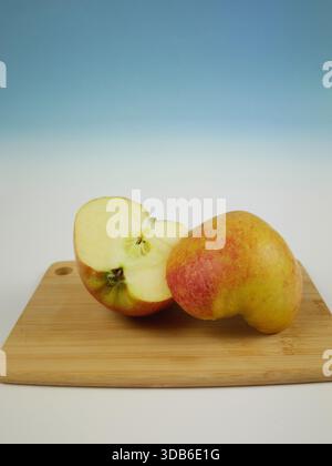 Vertikales Bild des ganzen Apfels und der Apfelhälfte auf Holzbrett vor weichem blauem Hintergrund, frische Fruchtzutaten für einfaches gebackenes Apfeldessert Stockfoto